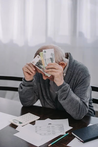 Stressed senior man sitting at table with paperwork and holding money at home — Stock Photo