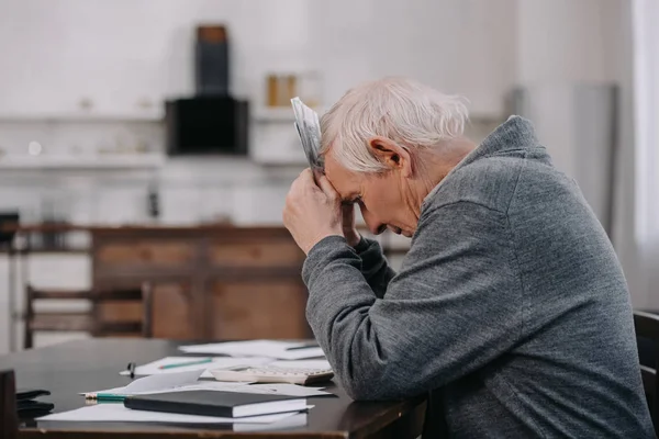 Gestresster Senior in Freizeitkleidung sitzt mit Papieren am Tisch und hält Geld zu Hause — Stockfoto