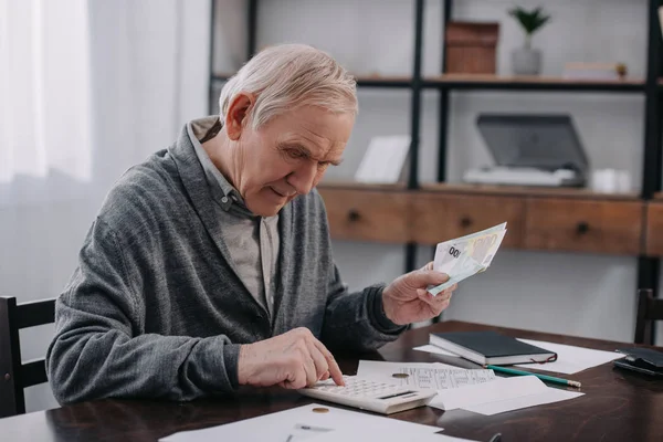Старший человек в повседневной одежде сидит за столом с бумажной работой и с помощью калькулятора при подсчете денег — стоковое фото