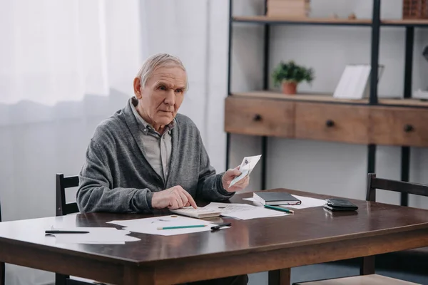 Senior man sitting at table with paperwork and using calculator while counting money at home — Stock Photo