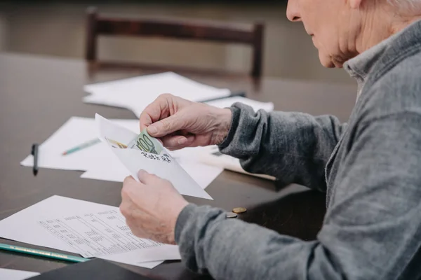 Обрезанный вид пожилого человека, сидящего за столом с бумажной работой и держащего конверт с деньгами — стоковое фото
