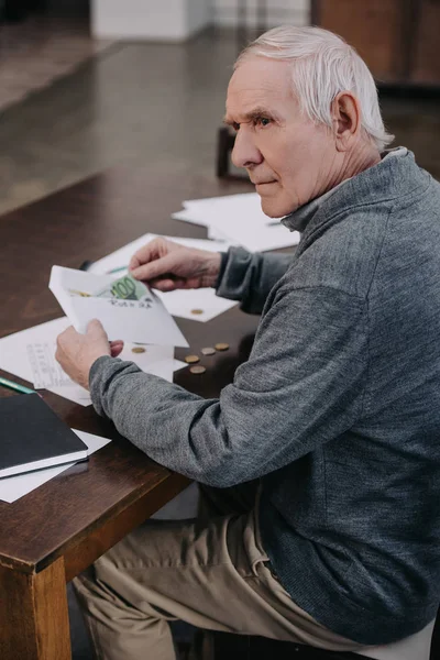 Senior assis à table avec de la paperasse et tenant enveloppe avec de l'argent — Photo de stock