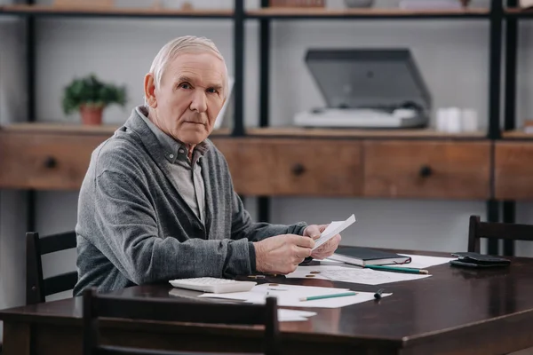 Senior man sitting at table with paperwork, money and holding envelope while looking at camera — Stock Photo