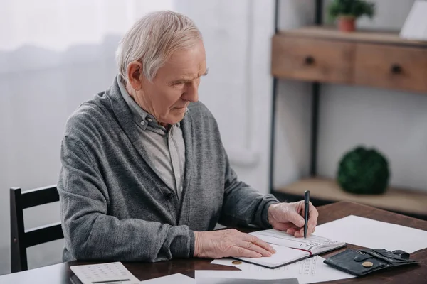 Senior sitzt mit Papierkram am Tisch und schreibt in Notizbuch — Stockfoto