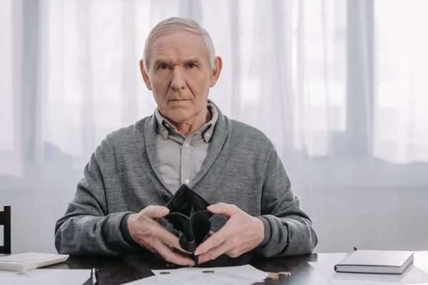 Pensionista masculino sentado en la mesa con papeleo, mirando a la cámara y sosteniendo la cartera vacía - foto de stock