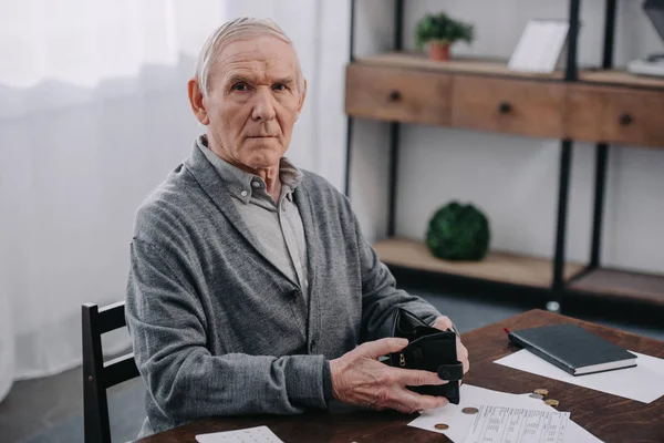 Чоловічий пенсіонер, сидячи за столом з документами, дивлячись на камеру і, тримаючи гаманець — стокове фото