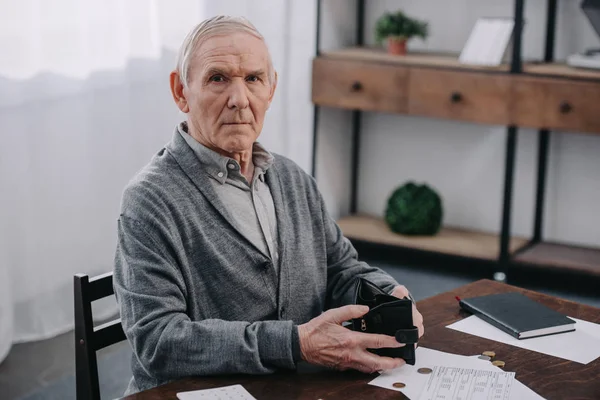 Pensionista masculino sentado en la mesa con papeleo, mirando a la cámara y sosteniendo la billetera — Stock Photo