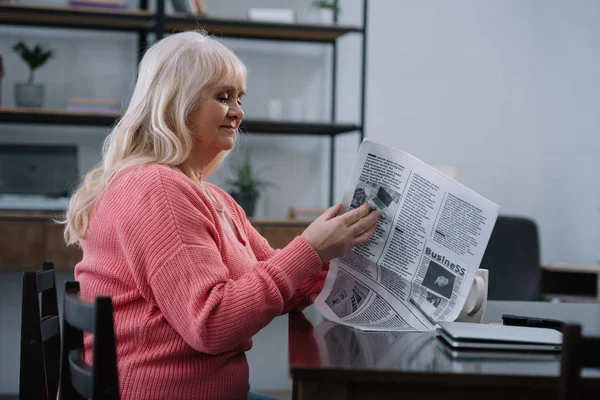 Старшая женщина сидит за столом и читает деловую газету дома — стоковое фото