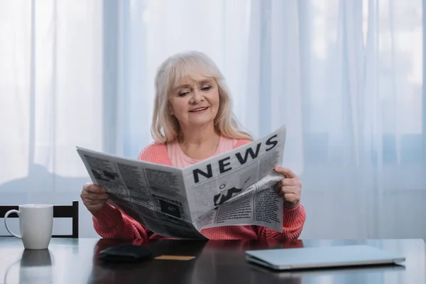 Lächelnde Seniorin sitzt zu Hause am Tisch und liest Zeitung — Stockfoto