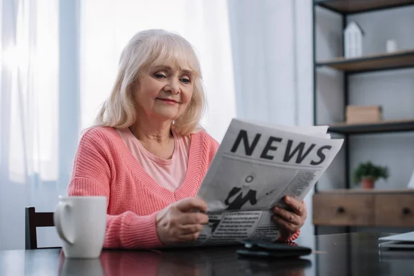 Улыбающаяся пожилая женщина, сидящая за столом и читающая газеты дома — стоковое фото