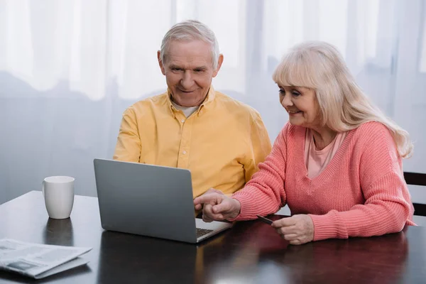 Sonriente pareja de ancianos en ropa casual usando el ordenador portátil mientras está sentado en la mesa en casa - foto de stock