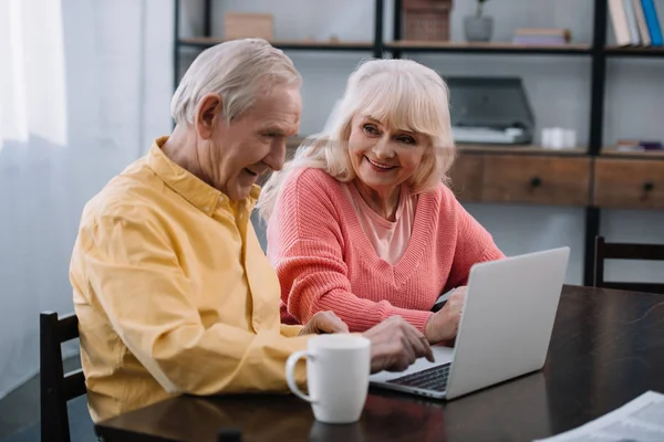 Smiling senior couple in casual clothes using laptop while sitting at table at home — Stock Photo