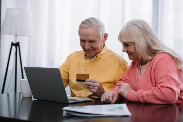 Пожилая пара, сидящая на диване с ноутбуком и кредитной картой во время покупок в Интернете дома — стоковое фото