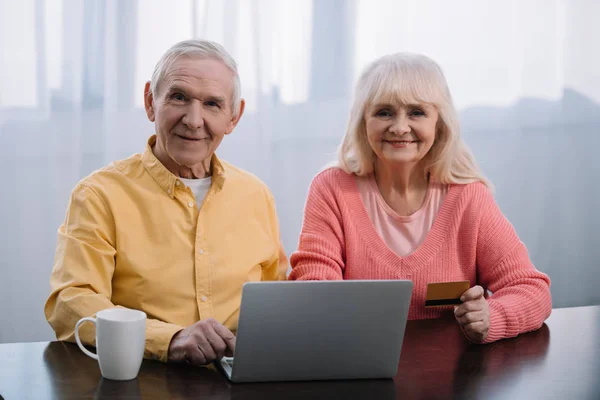 Casal idoso sentado no sofá com laptop e cartão de crédito, olhando para a câmera e fazendo compras on-line em casa — Fotografia de Stock