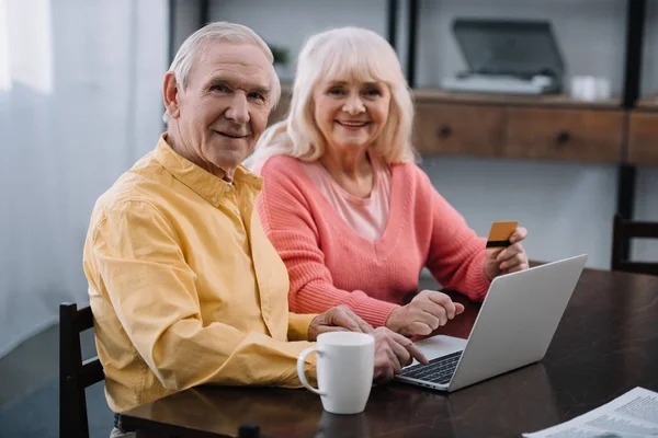 Seniorenpaar sitzt mit Laptop und Kreditkarte auf Couch, blickt in die Kamera und erledigt zu Hause Online-Einkäufe — Stockfoto