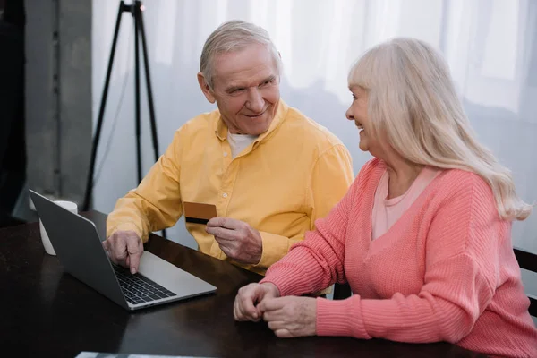 Счастливая пожилая пара сидит на диване с ноутбуком и кредитной картой, делая покупки в Интернете дома — стоковое фото