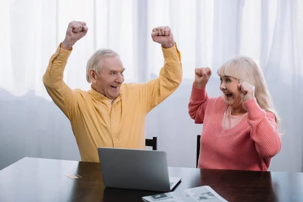 Happy senior couple in colorful clothes sitting at table with laptop and cheering with hands in air — Stock Photo