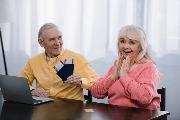 Senior hombre celebración de billetes de avión y pasaportes cerca de mujer sorprendida en casa - foto de stock