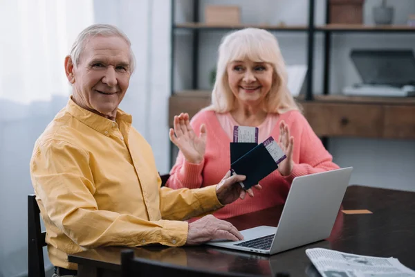 Старший чоловік дивиться на камеру і тримає авіаквитки з паспортами біля здивованої жінки вдома — стокове фото