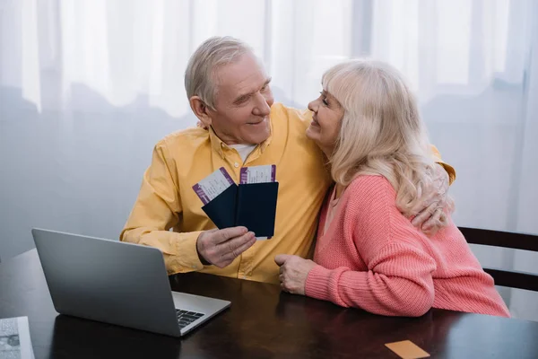 Улыбающаяся пожилая пара, сидящая за столом с ноутбуком, обнимающая и держащая авиабилеты с паспортами дома — стоковое фото