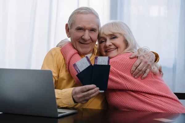 Heureux couple de personnes âgées assis à table avec ordinateur portable, étreignant et tenant des billets d'avion avec passeports à la maison — Photo de stock