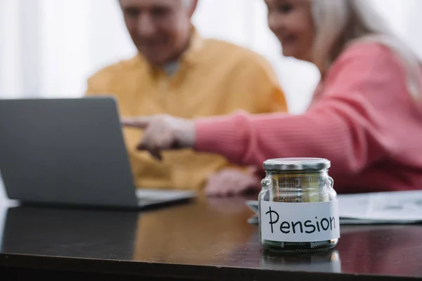 Selective focus of glass jar with 'pension' lettering and senior couple pointing with finger while using laptop on background — Stock Photo