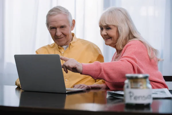 Sorridente coppia anziana seduta a tavola, che punta con il dito e utilizzando il computer portatile — Foto stock