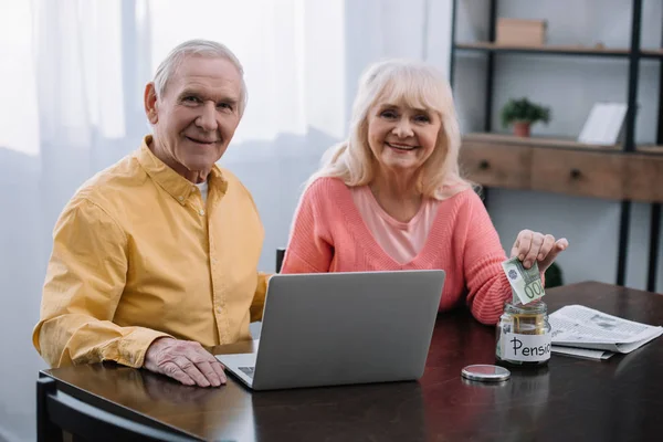 Pareja de ancianos sentados en la mesa con el ordenador portátil y mirando a la cámara, mientras que la mujer poniendo dinero en frasco de vidrio con letras 'pensión' - foto de stock