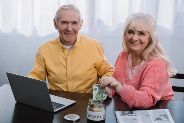 Pareja de ancianos sentados en la mesa con el ordenador portátil y mirando a la cámara, mientras que la mujer poniendo dinero en frasco de vidrio con letras 'pensión' - foto de stock