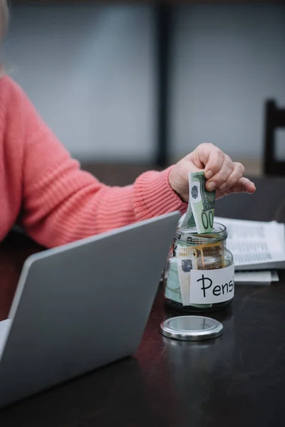 Обрезанный вид пожилой женщины, сидящей за столом с ноутбуком, в то время как положить деньги в стеклянную банку с 