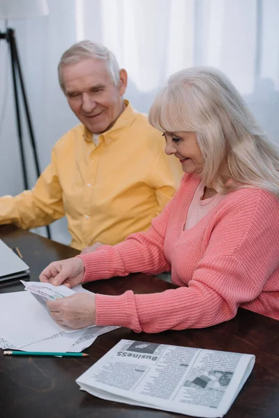 Lächelndes Seniorenpaar in bunten Kleidern sitzt mit Wirtschaftszeitung am Tisch — Stockfoto