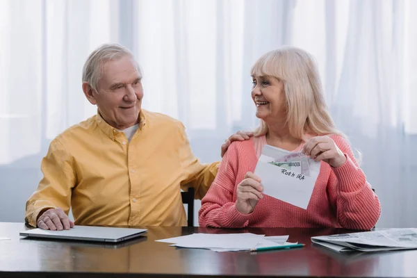 Pareja de ancianos sosteniendo sobre con letras 'roth ira' y dinero mientras están sentados en la mesa - foto de stock