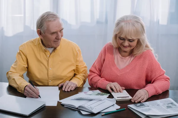 Lächelnder Senior sitzt mit Taschenrechner am Tisch und zählt Geld — Stockfoto