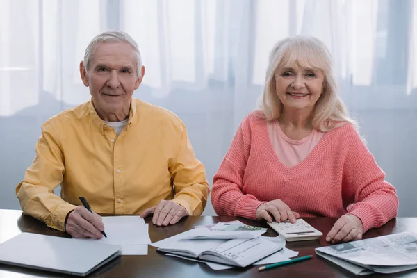 Senior coupe looking at camera while sitting at table with calculator and counting money — Stock Photo