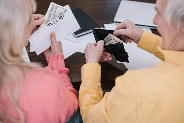 Visão traseira do casal sênior segurando dinheiro e envelope com letras 'roth ira' enquanto sentado na mesa — Fotografia de Stock