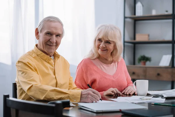 Щаслива старша пара сидить за столом і заповнює документи вдома — стокове фото