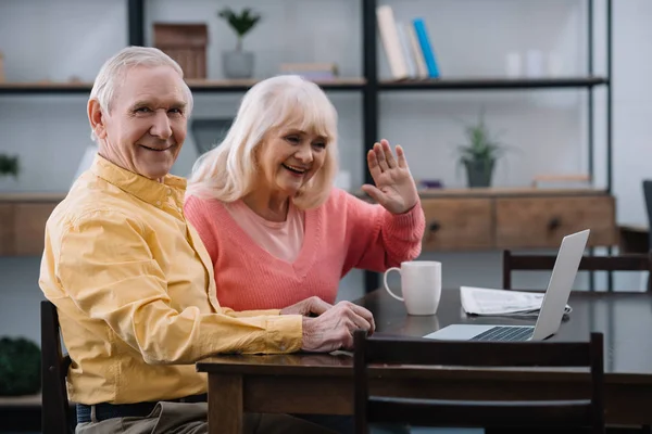 Sorridente coppia anziana seduta a tavola e utilizzando il computer portatile durante la videochiamata a casa — Foto stock