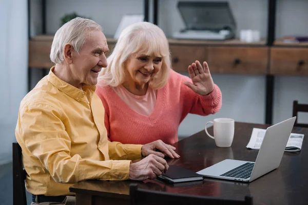 Lächelndes Senioren-Paar sitzt am Tisch und benutzt Laptop beim Videoanruf zu Hause — Stockfoto