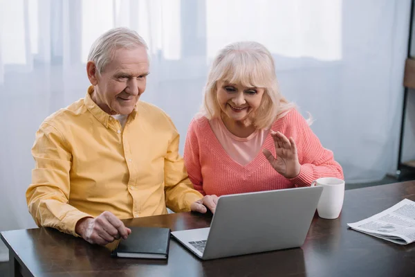 Pareja de ancianos sentados en la mesa y el uso de ordenador portátil durante la videollamada en casa - foto de stock