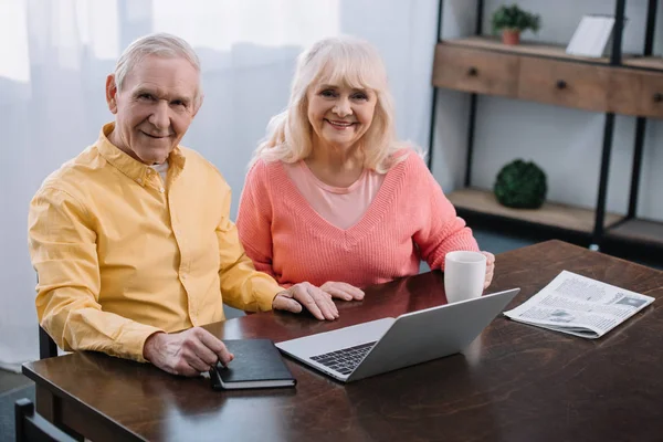 Lächelndes Seniorenpaar blickt in die Kamera und benutzt Laptop, während es zu Hause am Tisch sitzt — Stockfoto