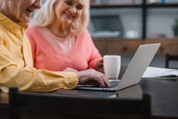 Sourire couple aîné en vêtements colorés assis à la table et en utilisant un ordinateur portable — Photo de stock