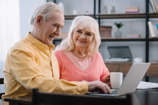 Heureux couple âgé en vêtements colorés assis à la table et en utilisant un ordinateur portable — Photo de stock