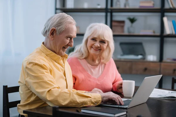 Pareja de ancianos en ropa colorida sentado en la mesa con taza de café y el uso de ordenador portátil - foto de stock