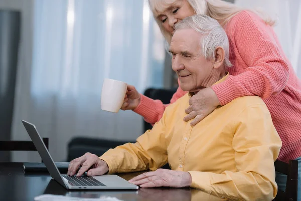 Seniorin hält Kaffeetasse und umarmt Mann, der am Tisch sitzt und Laptop benutzt — Stockfoto