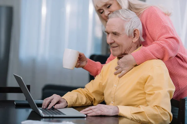 Senior femme tenant tasse de café et câlin homme assis à la table et en utilisant un ordinateur portable — Photo de stock