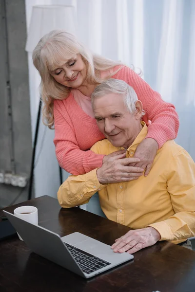Sourire couple aîné étreignant tandis que l'homme assis à la table et en utilisant un ordinateur portable — Photo de stock