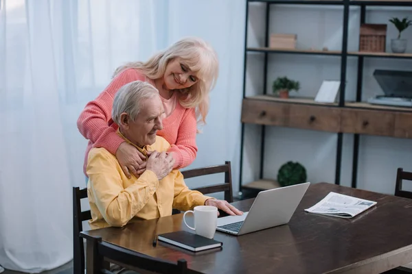 Glückliches Senioren-Paar umarmt, während Mann am Tisch sitzt und Laptop im Wohnzimmer benutzt — Stockfoto