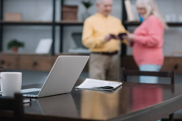 Selective focus of laptop and newspaper on table with senior couple on background — Stock Photo