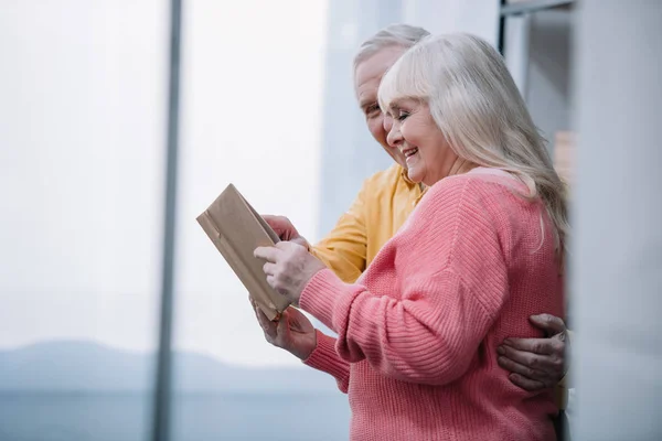 Heureux couple aîné en vêtements colorés livre de lecture à la maison avec espace de copie — Photo de stock
