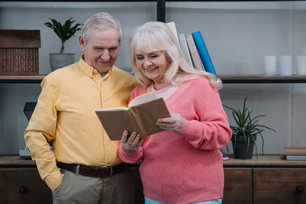 Glückliches Seniorenpaar in bunten Kleidern liest zu Hause Buch — Stockfoto